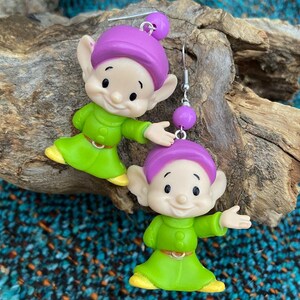 Può includere: Un paio di orecchini pendenti con un personaggio dei cartoni animati con capelli viola, vestiti verdi e un cappello giallo. Il personaggio sorride e ha grandi orecchie. Gli orecchini sono fatti di plastica e hanno ganci d'argento.