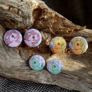 Può includere: Sei bottoni rotondi con disegni di pony dei cartoni animati in rosa, giallo e verde. Ogni bottone ha uno sfondo a scacchi. I bottoni sono disposti su un pezzo di legno trasportato dalle correnti.