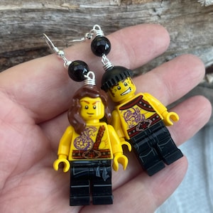 Könnte beinhalten: Ein Paar Ohrringe mit Lego-Minifiguren mit schwarzen und violetten Tattoos, die schwarze Hosen und einen schwarzen Hut tragen. Die Ohrringe sind aus schwarzen Perlen und Silberdraht gefertigt.