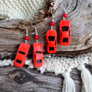 May include: Bright red car-shaped earrings with black windows and silver hooks. Each earring features a small red bead. The earrings are displayed on a piece of driftwood, with a cream-colored knitted background.
