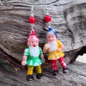 May include: A pair of whimsical earrings featuring miniature gnome figures. One gnome wears a green tunic and yellow pants, the other a yellow tunic and red pants. Each earring has a red bead and silver-tone ear wire.