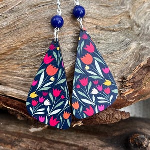 Può includere: Un paio di orecchini pendenti con sfondo blu scuro e un motivo floreale con tulipani rosa, arancioni, gialli e bianchi. Gli orecchini hanno una perla blu scuro nella parte superiore del gancio argentato.
