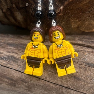 Könnte beinhalten: Ein Paar Ohrringe mit Lego-Minifiguren von Höhlenmenschen mit braunen Haaren, gelber Haut und braunen Lendenschürzen. Die Minifiguren sind an silbernen Ohrhaken mit schwarzen Perlen befestigt.