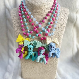 Può includere: Una collana multistrato con perline color azzurro pastello, rosa e verde. Ogni strato ha un piccolo ciondolo a forma di pony di plastica con un fiocco. I pony sono rosa, blu, giallo e viola.