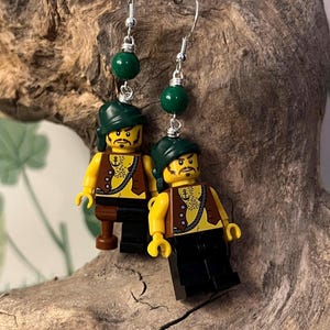 Puede incluir: Un par de pendientes con minifiguras Lego vestidas de piratas. Cada minifigura lleva un chaleco marrón, pantalones negros y un sombrero verde. Los pendientes están adornados con cuentas verdes.