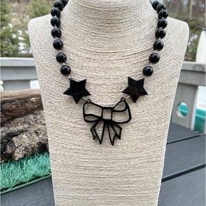 Può includere: Una collana nera con un grande ciondolo a forma di fiocco nero e due ciondoli a forma di stella nera. La collana è composta da perle rotonde nere.