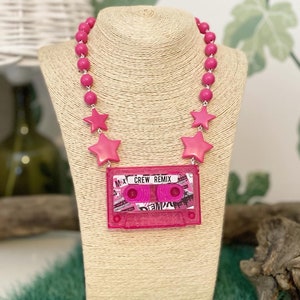 Può includere: Una collana di perline rosa con ciondoli a forma di stella e un ciondolo a forma di cassetta rosa che dice "CREW REMIX".