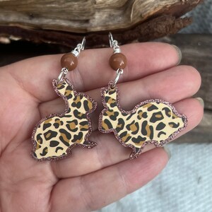 Può includere: Un paio di orecchini pendenti a forma di coniglietto con un motivo leopardato. Gli orecchini hanno un bordo glitterato color oro rosa e sono sospesi da ganci argentati con un'accentuazione di perline marroni.