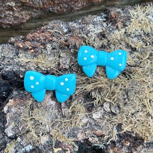 Può includere: Due accessori a forma di fiocco azzurro chiaro con pois bianchi. I fiocchi sono posizionati su una superficie marrone strutturata con muschio. Gli accessori sono probabilmente orecchini o fermagli per capelli.