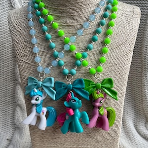 Può includere: Tre collane colorate con ciondoli a forma di pony. Ogni collana ha perline di un colore diverso: azzurro, turchese e verde neon. I ciondoli a forma di pony sono attaccati a fiocchi di colori abbinati.