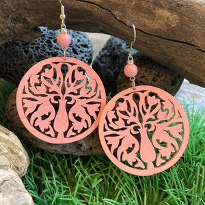 Può includere: Un paio di orecchini in legno tagliati al laser color corallo. Ogni orecchino presenta un disegno ad albero all'interno di una cornice circolare, con una piccola perla rosa sopra. Gli orecchini pendono da fili argentati.