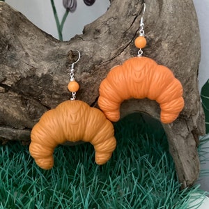 Può includere: Un paio di orecchini a forma di croissant arancioni con ganci e perline argentate. Gli orecchini sono appesi a un pezzo di legno galleggiante.