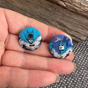 Può includere: Due bottoni rotondi in tessuto di Batman. Un bottone raffigura Batman con mantello e cappuccio blu, con un simbolo del pipistrello giallo. L'altro bottone mostra Batman che lancia un batarang. I bottoni sono su una superficie strutturata.