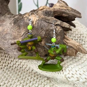 Könnte beinhalten: Handgefertigte Ohrringe mit grünen Miniatur-Teenage Mutant Ninja Turtles-Figuren. Jeder Ohrring hat eine grüne Perle und einen silberfarbenen Ohrdraht. Die Schildkröten posieren mit ihren Waffen, bereit für die Action.
