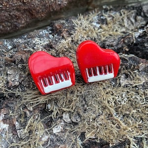 Può includere: Due orecchini rossi a forma di cuore con tasti di pianoforte bianchi. Gli orecchini sono su uno sfondo di legno strutturato con muschio.