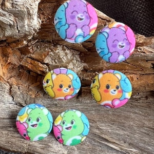 Può includere: Sei bottoni rotondi rivestiti in tessuto con disegni di orsetti dei cartoni animati. I bottoni presentano orsetti viola, arancioni e verdi con accenti blu e rosa. Sembrano fatti a mano e sono su una superficie di legno.