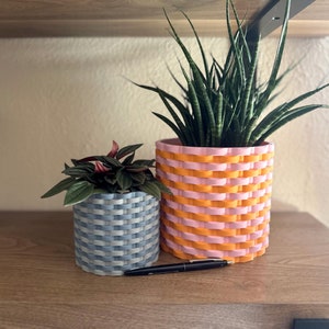 May include: Two decorative woven-style planters. One is blue and grey, the other is pink and orange. Both planters contain green plants. A black pen rests on the wooden surface in front of the planters. The planters are a modern home decor item.