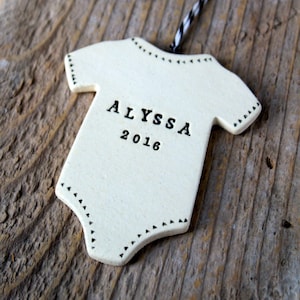 May include: White ceramic ornament shaped like a baby onesie with black stitching and the name "ALYSSA" and the year "2016" printed on it. The ornament is hanging from a black and white striped string.
