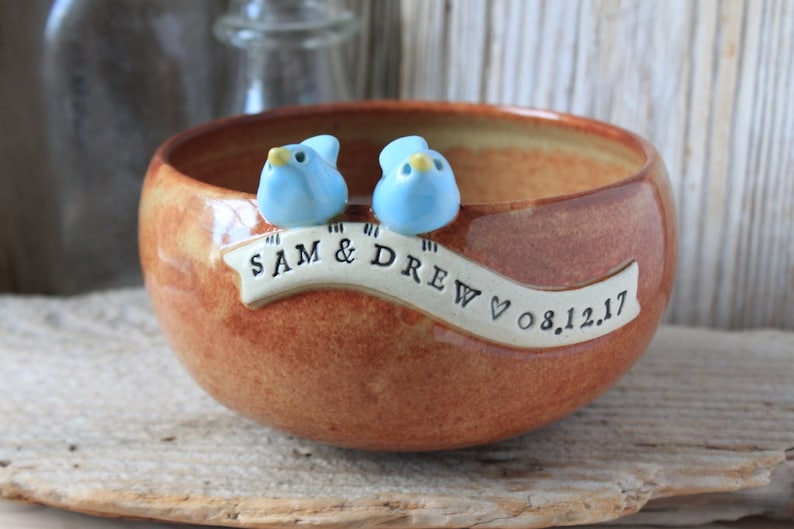 May include: A brown ceramic bowl with two blue bird figurines on the rim. The bowl has a white banner with the text "SAM & DREW" and the date "08.12.17".