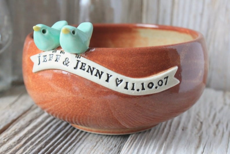Pode incluir: Uma tigela de cer&acirc;mica marrom com dois p&aacute;ssaros azul claro na borda. A tigela tem uma faixa branca com o texto "JEFF & JENNY &hearts; 11.10.07"