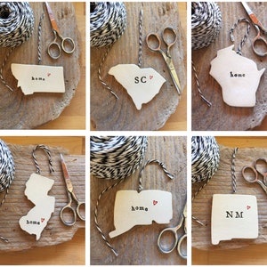 May include: Six white ceramic ornaments shaped like different states in the US. Each ornament has a red heart and text on it. The text includes the state abbreviation and the word "home".