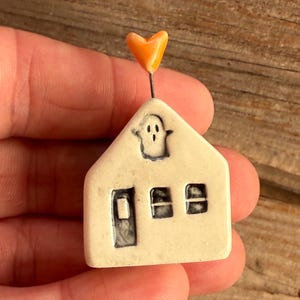 May include: A small, off-white ceramic house ornament featuring a dark-outlined ghost in the attic window, a door, and two windows. A thin metal wire extends from the roof, topped with a small, glossy, orange heart-shaped accent piece.