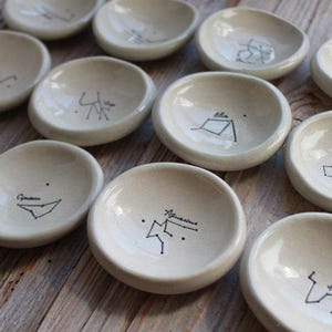 May include: Twelve small, white ceramic dishes with black constellation drawings. Each dish features a different constellation with its name written below.