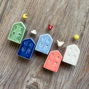 May include: Five miniature ceramic houses in different colors, each with a unique detail on top. The houses are green, blue, light blue, pink, and white. The details on top are a crescent moon, a cloud, a heart, a bird, and a sun.