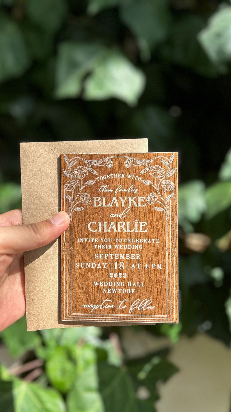 Pu&ograve; includere: Un invito di nozze in legno con decorazioni floreali e i nomi Blayke e Charlie. L'invito include la data, l'ora e il luogo del matrimonio a New York.