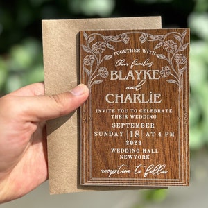 Pu&ograve; includere: Un invito di nozze in legno con dettagli floreali e i nomi "Blayke e Charlie" incisi in bianco. L'invito include la data, l'ora e il luogo del matrimonio. Tenuto con una busta marrone.