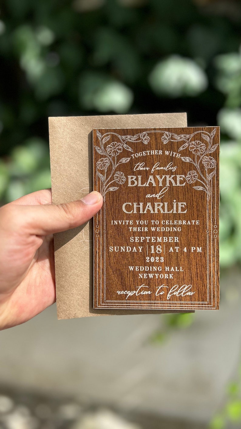 Pu&ograve; includere: Un invito di nozze in legno con dettagli floreali e i nomi "Blayke e Charlie" incisi in bianco. L'invito include la data, l'ora e il luogo del matrimonio. Tenuto con una busta marrone.