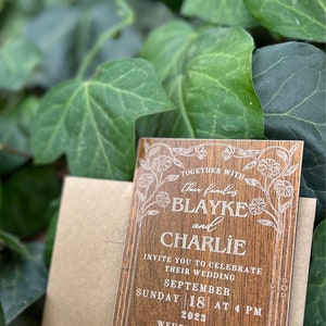 Pu&ograve; includere: Un invito di nozze in legno con dettagli floreali e i nomi "Blayke e Charlie" in elegante scrittura. L'invito include la data, l'ora e il luogo del matrimonio, con una busta abbinata. Lo sfondo presenta foglie verdi.