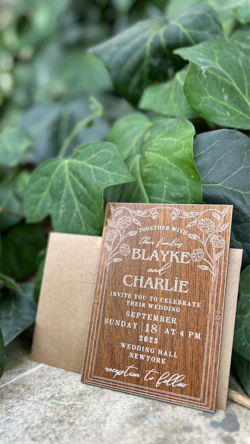 Pu&ograve; includere: Un invito di nozze in legno con dettagli floreali e i nomi "Blayke e Charlie" in elegante scrittura. L'invito include la data, l'ora e il luogo del matrimonio, con una busta abbinata. Lo sfondo presenta foglie verdi.