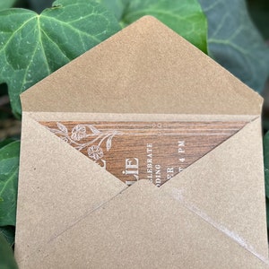 Pu&ograve; includere: Una busta di carta kraft parzialmente aperta, che rivela un invito con texture di legno. L'invito presenta delicati disegni floreali e il testo "Celebrate Wedding" e "4 PM". Lo sfondo &egrave; un fogliame verde sfocato.