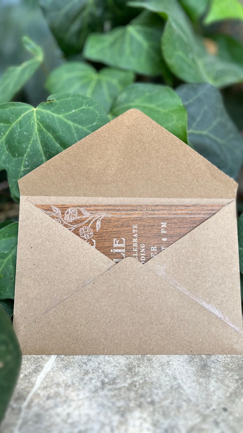 Pu&ograve; includere: Una busta di carta kraft parzialmente aperta, che rivela un invito con texture di legno. L'invito presenta delicati disegni floreali e il testo "Celebrate Wedding" e "4 PM". Lo sfondo &egrave; un fogliame verde sfocato.