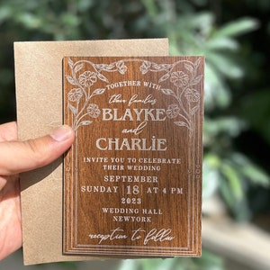 Pu&ograve; includere: Un invito di nozze in legno con dettagli floreali incisi e i nomi "Blayke e Charlie". L'invito include la data, l'ora e il luogo del matrimonio. &Egrave; tenuto con una busta marrone.