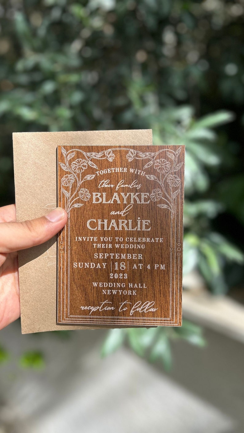 Pu&ograve; includere: Un invito di nozze in legno con dettagli floreali incisi e i nomi "Blayke e Charlie". L'invito include la data, l'ora e il luogo del matrimonio. &Egrave; tenuto con una busta marrone.