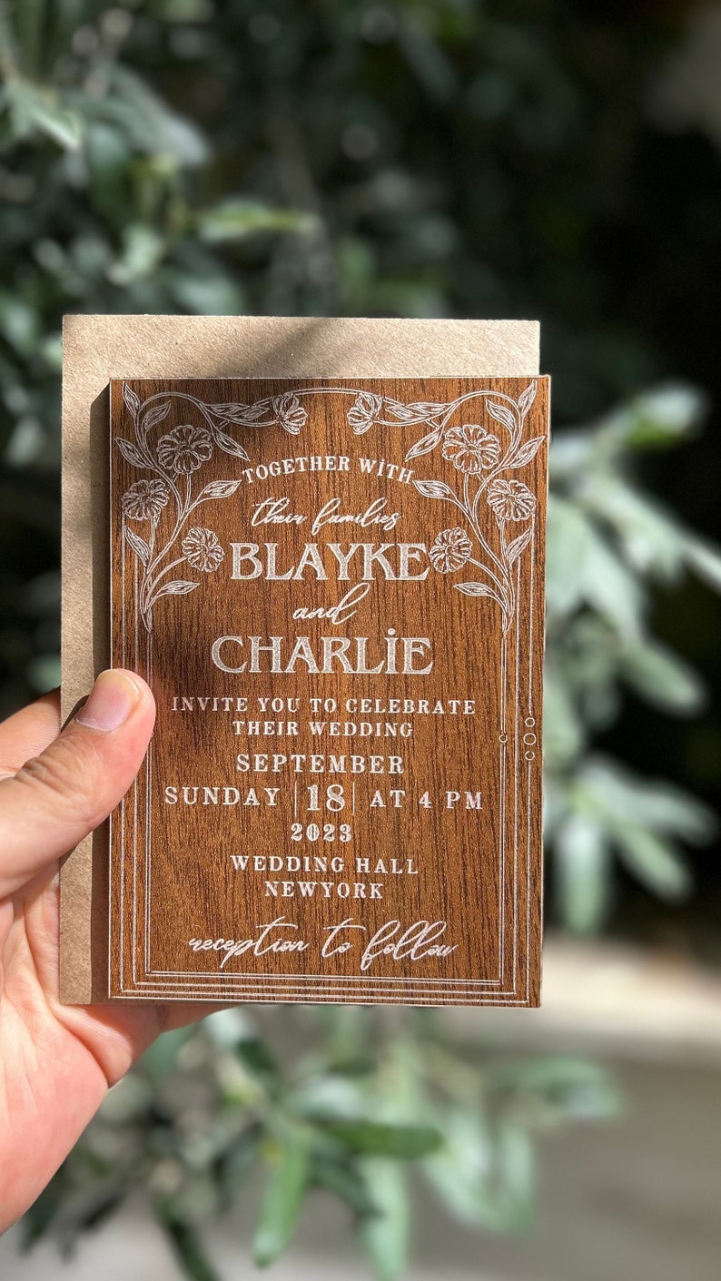 Pu&ograve; includere: Un invito di nozze in legno con testo inciso e dettagli floreali. L'invito recita "Blayke e Charlie" e include la data, l'ora e il luogo del matrimonio. L'invito &egrave; tenuto con una busta di carta kraft.