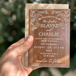 Pu&ograve; includere: Un invito di nozze in legno con dettagli floreali incisi e i nomi "Blayke e Charlie". L'invito include la data, l'ora e il luogo del matrimonio. &Egrave; tenuto davanti a uno sfondo verde sfocato.