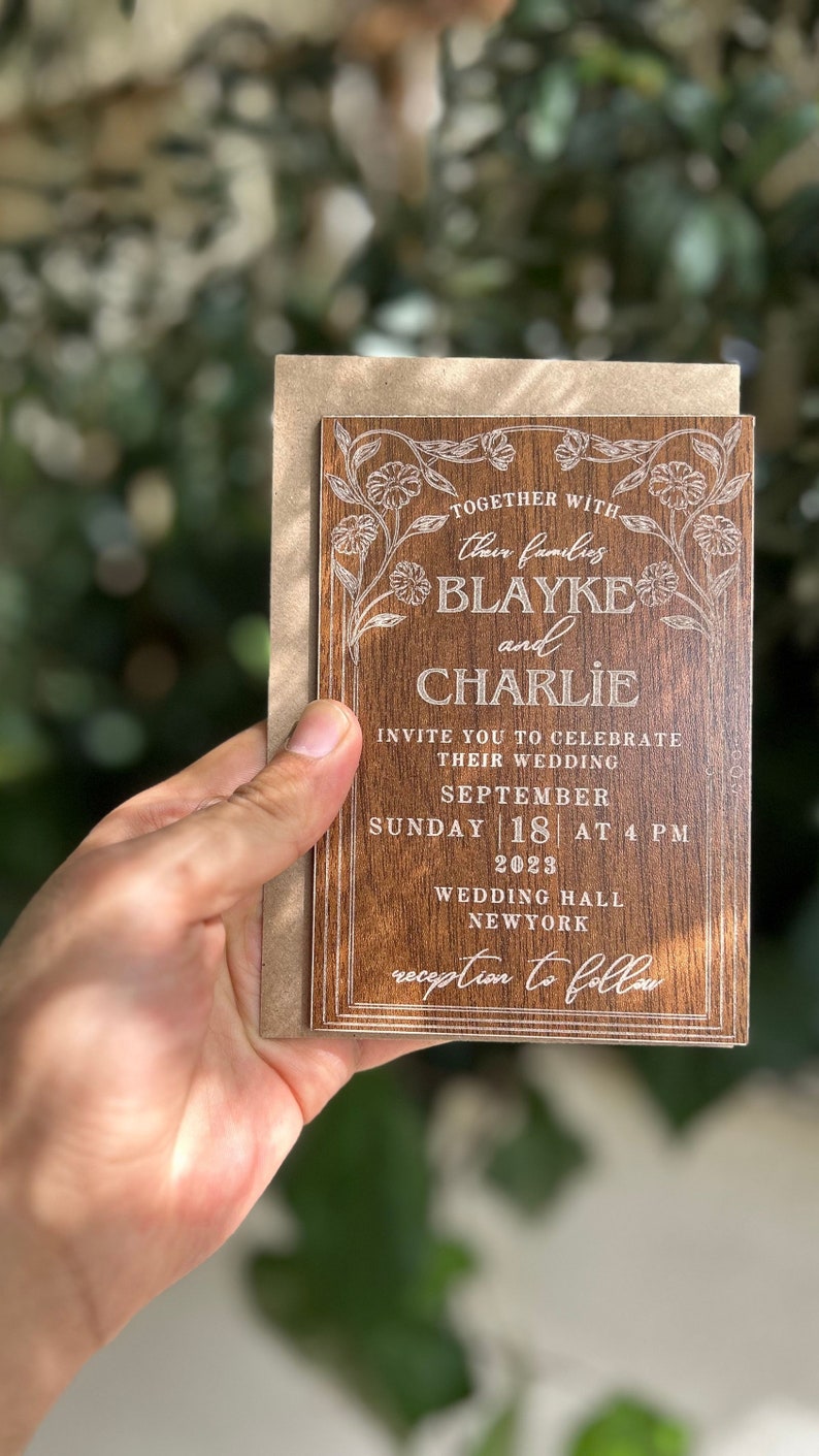 Pu&ograve; includere: Un invito di nozze in legno con dettagli floreali incisi e i nomi "Blayke e Charlie". L'invito include la data, l'ora e il luogo del matrimonio. &Egrave; tenuto davanti a uno sfondo verde sfocato.