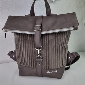 Könnte beinhalten: Ein grauer Cordrucksack mit Klappenverschluss und silbernem Reißverschluss. Der Rucksack hat eine silberne Schnalle und einen silbernen Riemen. Der Rucksack trägt den Schriftzug "Handmade" auf der Vorderseite.