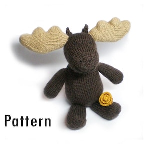 May include: A brown knitted moose toy with tan antlers and a yellow flower on its leg. The toy is sitting on a white background.
