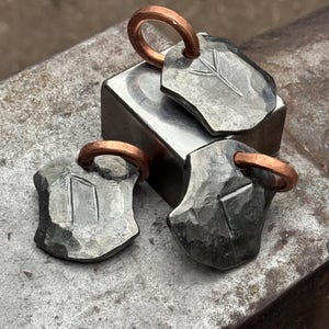 Może przedstawiać: Trzy srebrne metalowe zawieszki z miedzianymi kółkami. Każda zawieszka ma kształt tarczy z wygrawerowanym innym symbolem runicznym.