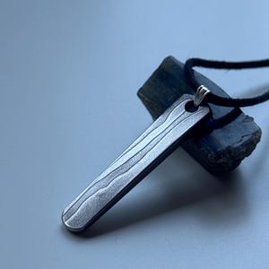 Könnte beinhalten: Ein silberfarbener rechteckiger Anhänger mit Holzmaserung hängt an einer schwarzen Kordel. Der Anhänger liegt auf einem dunkelgrauen Stein vor einem hellgrauen Hintergrund. Der Anhänger hat einen abgerundeten Boden und eine kleine Schlaufe oben für die Kordel.