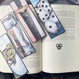 以下が含まれることがあります： 水彩画のイラストが描かれた、異なる4つのしおり。1つのしおりには剣が、もう1つのしおりには蛇が、もう1つのしおりにはサイコロのセットが、最後のしおりには3つの円が描かれたシンボルが描かれています。