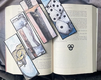 May include: Four different bookmarks with watercolor illustrations. One bookmark features a sword, another a snake, another a set of dice, and the last one has a symbol of three circles.