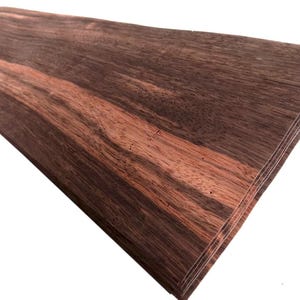 May include: A stack of thin, rectangular wood veneer sheets featuring a prominent grain pattern with alternating dark brown and reddish-brown stripes. The sheets are layered together, displaying a smooth, flat surface with visible wood fibers.