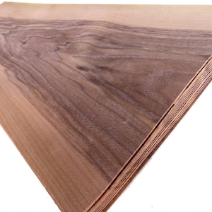 May include: A stack of wood veneer sheets. The top sheet displays a natural wood grain pattern with shades of brown and tan. The edges of the sheets are visible, showing the layered construction. The wood has a smooth, polished appearance.