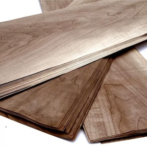 May include: A stack of thin, light brown wood veneer sheets with visible grain patterns. The sheets are arranged in a layered fashion, with some fanned out to display the wood's texture. The wood has a natural, warm tone.