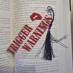 May include: A close-up of a blue tassel bookmark with a red and white glitter design. The bookmark is on a page of a book with text about magic and spells. The text on the page reads "There was no real magic at this mark nor lore."